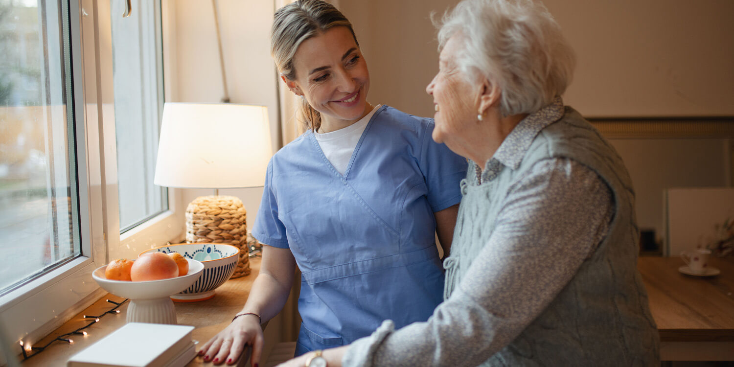 Caregiver in blue scrubs smiling with an older woman at home