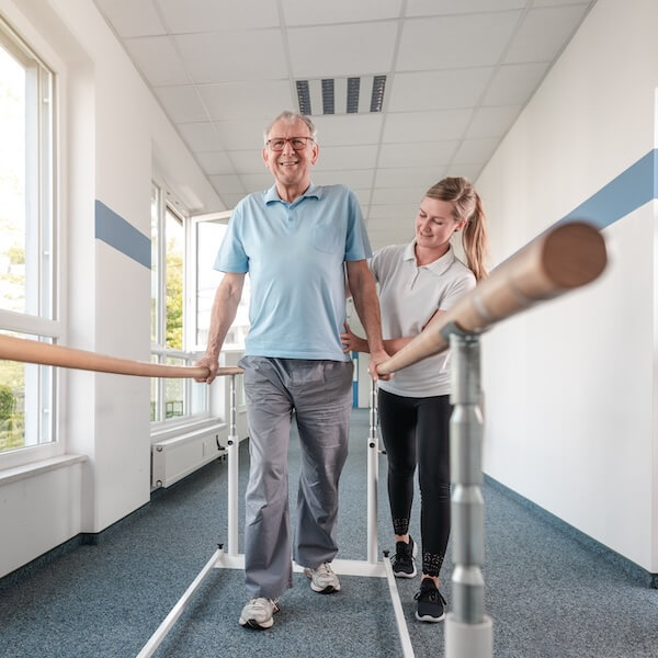 Physical therapist helping a senior man walk with parallel bars in a hallway